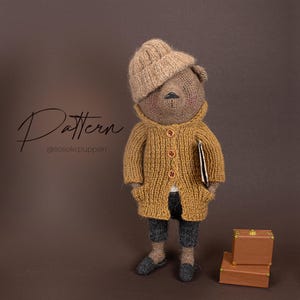 Pode incluir: Um urso de peluche de malha marrom vestindo um chapéu de malha marrom, um casaco de malha amarelo e calças pretas. O urso está segurando um livro marrom e está em pé na frente de duas malas marrons.