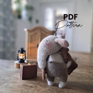 Könnte beinhalten: Ein gestricktes Elefantenspielzeug mit Rüssel, Ohren und einem braunen Schal. Der Elefant trägt eine braune Latzhose und hat einen kleinen Koffer. Der Text "PDF Pattern" ist kursiv geschrieben. Das Spielzeug steht auf einer Holzoberfläche.