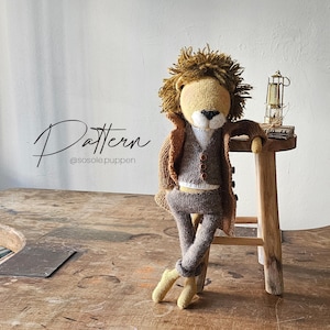 Puede incluir: Un león de crochet que lleva una chaqueta marrón y pantalones grises. El león está de pie sobre un taburete de madera con una linterna y un libro en la parte superior. El texto "Pattern @sosole.puppen" es visible en la imagen.
