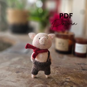 May include: A knitted piglet doll wearing brown overalls and a red scarf. The piglet has pink ears and cheeks, and is standing upright. The image also includes the text "PDF Pattern".