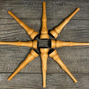 May include: Eight wooden bottle stoppers are arranged in a starburst design. Each stopper features a light brown, turned wood body and a black base. The wood grain is visible. The stoppers are displayed on a dark wood surface.