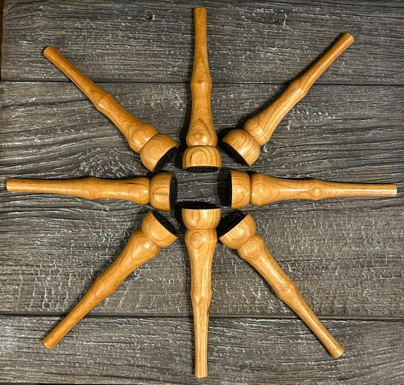 May include: Eight wooden bottle stoppers are arranged in a starburst design. Each stopper features a light brown, turned wood body and a black base. The wood grain is visible. The stoppers are displayed on a dark wood surface.