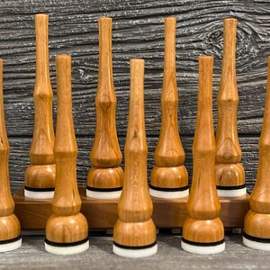 May include: Nine wooden bottle stoppers with a light brown finish. Each stopper has a unique shape with a rounded base, a cylindrical body, and a tapered top. The bases have a white and black ring. They are displayed on a wooden stand.
