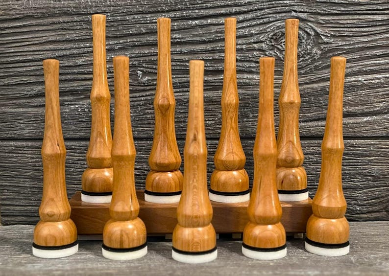 May include: Nine wooden bottle stoppers with a light brown finish. Each stopper has a unique shape with a rounded base, a cylindrical body, and a tapered top. The bases have a white and black ring. They are displayed on a wooden stand.