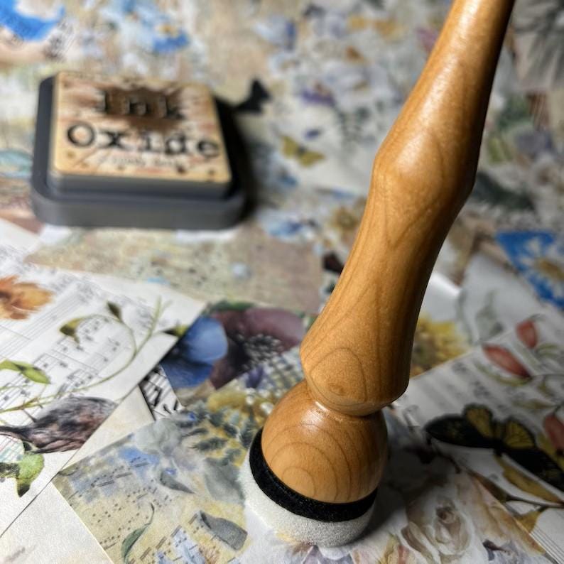 May include: A wooden handled blending tool with a white foam applicator and a black band. The tool is resting on a surface with patterned papers and an ink pad that reads "Ink Oxide".