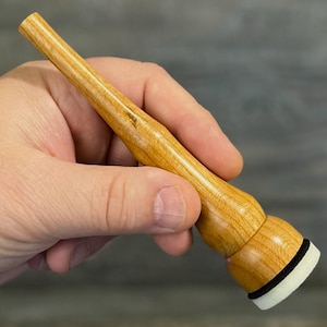 May include: A light-coloured wooden tool with a rounded base and a tapered handle. The base has a black ring and a white, cylindrical tip. The tool is held in a hand against a blurred background.
