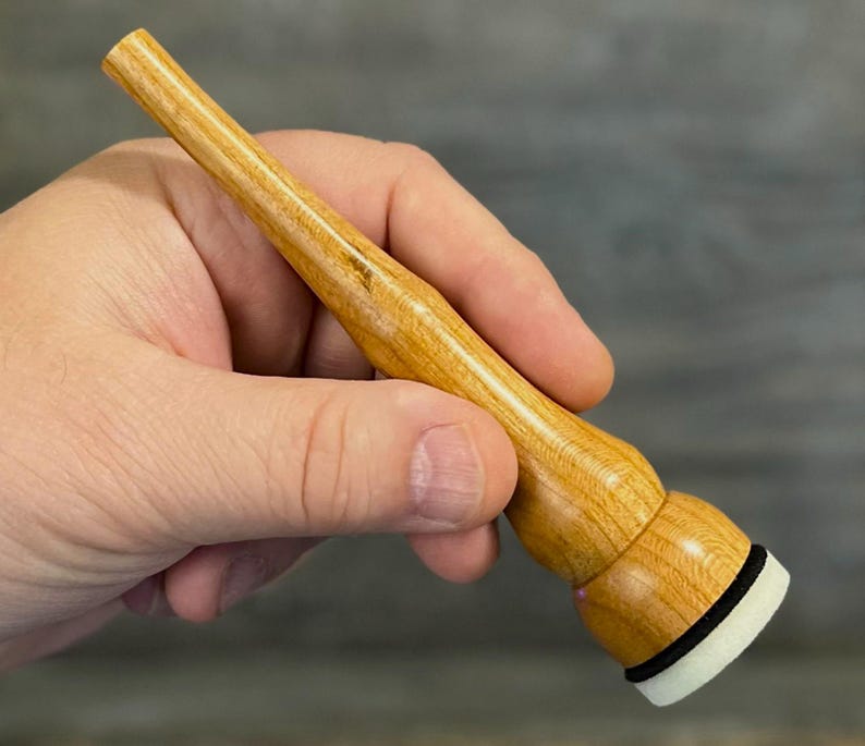 May include: A light-coloured wooden tool with a rounded base and a tapered handle. The base has a black ring and a white, cylindrical tip. The tool is held in a hand against a blurred background.
