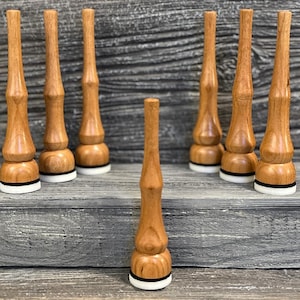 May include: A selection of wooden bottle stoppers. The stoppers are light brown with a rounded base, a black band, and a white ring. The stoppers are displayed on a grey wooden surface.