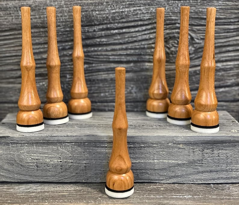 May include: A selection of wooden bottle stoppers. The stoppers are light brown with a rounded base, a black band, and a white ring. The stoppers are displayed on a grey wooden surface.