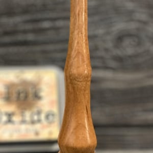 May include: A wooden stamp with a light brown finish. The stamp has a long, slender handle and a rounded base with a white and black pad. The stamp is designed for applying ink to paper or other surfaces.