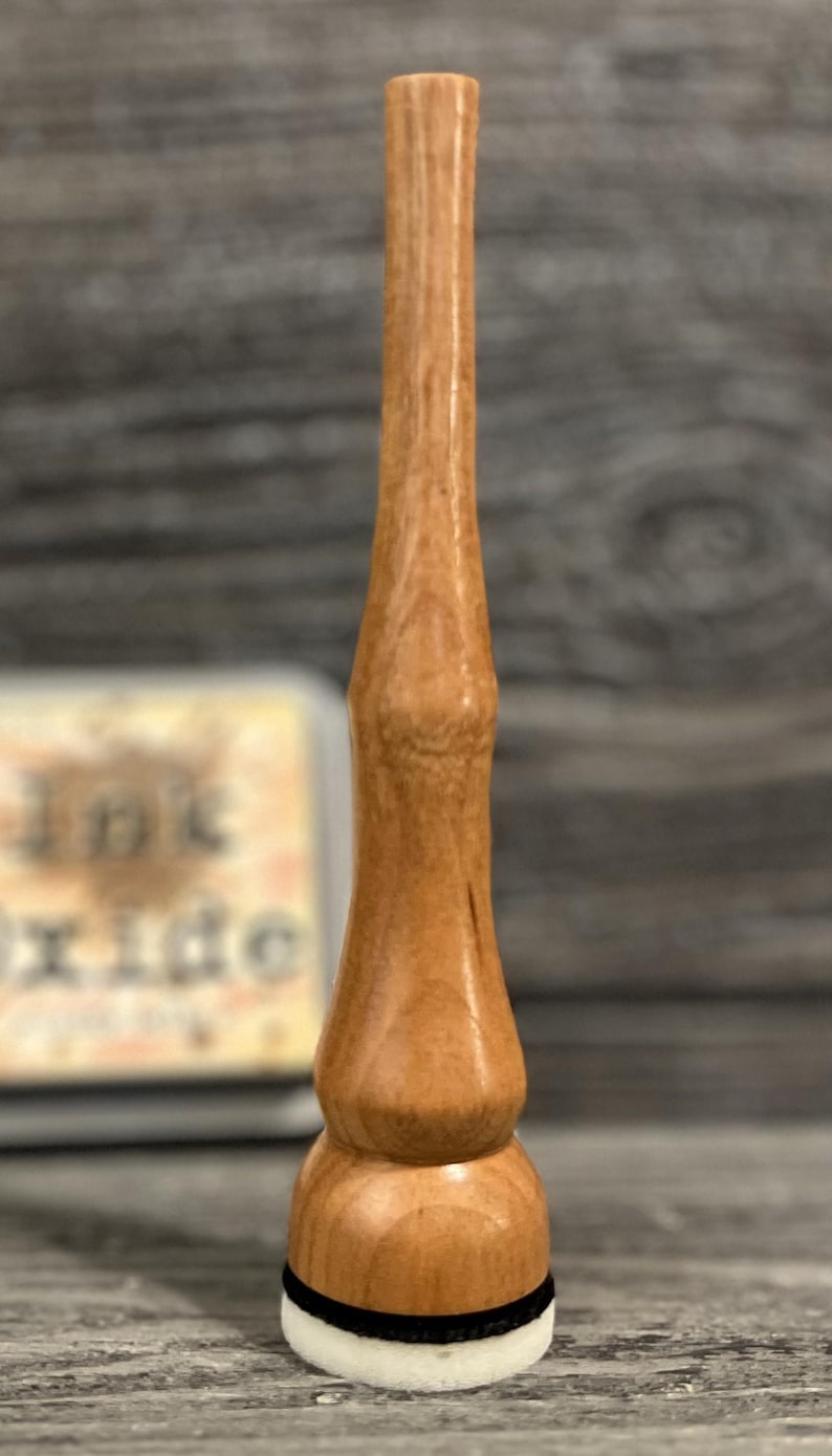 May include: A wooden stamp with a light brown finish. The stamp has a long, slender handle and a rounded base with a white and black pad. The stamp is designed for applying ink to paper or other surfaces.