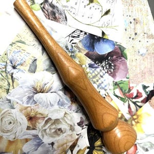 May include: A light brown wooden rolling pin. The handle is long and cylindrical, with a bulbous end. The rolling pin is made of light-coloured wood and is designed for rolling out dough. The rolling pin measures approximately 30 cm in length.