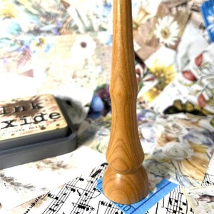 May include: A light brown wooden stamp handle with a rounded base and a slender, curved shaft. The handle is made of wood and has a natural grain pattern. The background includes a gray ink pad and a collage of floral and musical note papers.