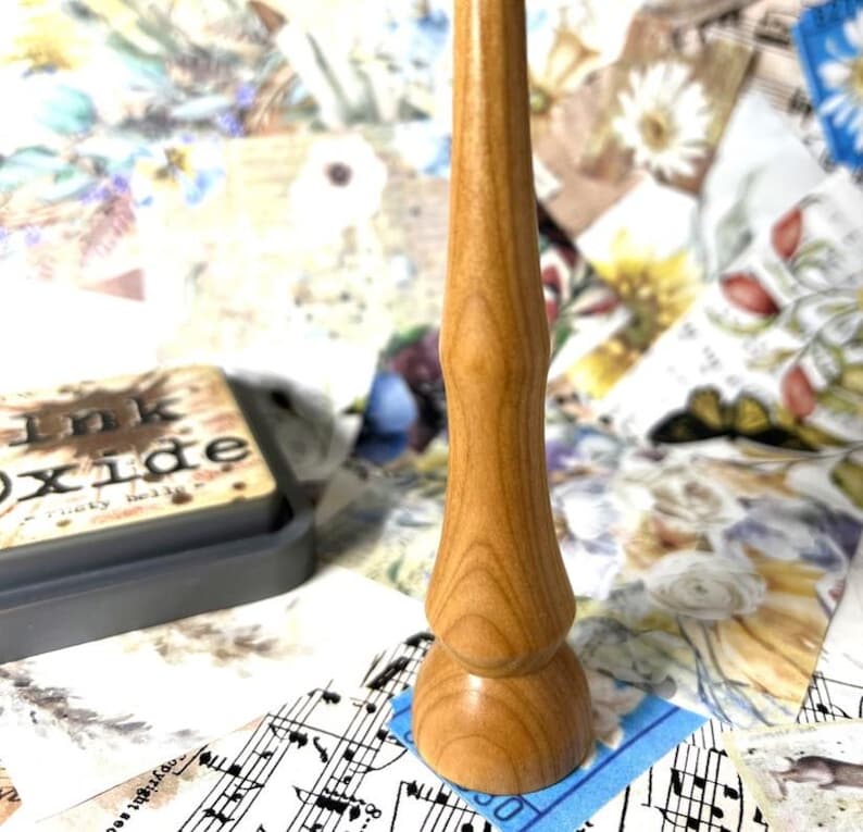 May include: A light brown wooden stamp handle with a rounded base and a slender, curved shaft. The handle is made of wood and has a natural grain pattern. The background includes a gray ink pad and a collage of floral and musical note papers.