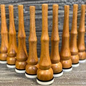 May include: Nine turned wooden bottle stoppers are displayed. Each stopper has a light brown finish, a bulbous base, a cylindrical midsection, and a tapered top. The base features a white and black ring. The stoppers are arranged in a triangular pattern.