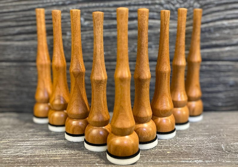 May include: Nine turned wooden bottle stoppers are displayed. Each stopper has a light brown finish, a bulbous base, a cylindrical midsection, and a tapered top. The base features a white and black ring. The stoppers are arranged in a triangular pattern.