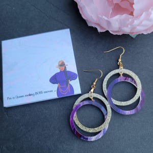 May include: Purple and silver glitter hoop earrings with a gold hook. The earrings have a layered design with a smaller circle inside a larger circle.