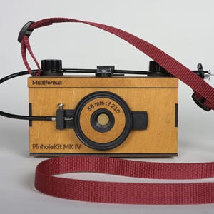 Könnte beinhalten: Eine Holz-Lochkamera mit einem weinroten Riemen. Die Kamera hat ein schwarzes Objektiv und ein schwarzes Kabel. Der Text "Multiformat" und "PinholeKit MK IV" sind auf der Kamera aufgedruckt.