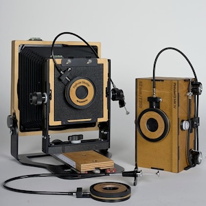 Könnte beinhalten: Zwei Lochkameras werden gezeigt. Die größere Kamera hat einen Holzrahmen, einen schwarzen Balgen und die Aufschrift "Pinhole Shutter". Die kleinere Kamera ist eine Box mit einer kreisförmigen Öffnung und dem Text "PinholeKit MK IV". Beide haben schwarze Kabel.