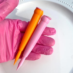 May include: Two colorful cosmetic applicators. One is bright orange, the other is pink. Both have a cylindrical body and a pointed tip. The applicators are held in a pink gloved hand, against a white background.