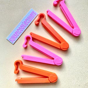 Könnte beinhalten: Fünf orangefarbene und rosafarbene Plastik-Ausstecher. Die Ausstecher sind herzförmig und haben einen kleinen Griff. Der Text "SUNSETCOOKIECUTTERS" ist im Hintergrund sichtbar.