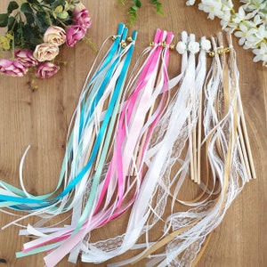 Può includere: Assortimento di bacchette di nastro colorate con bastoncini di legno e dettagli dorati. I nastri sono in tonalità di blu, verde, rosa e bianco. Alcuni nastri sono in raso e altri in pizzo.