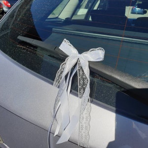 Puede incluir: Un lazo blanco de encaje atado al limpiaparabrisas trasero de un coche plateado. El lazo está hecho de cinta de satén blanco y encaje.