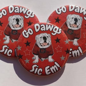 May include: Three red circular buttons with a bulldog graphic wearing a red jersey. The buttons have the text "Go Dawgs" and "Sic Em!" in white. Black stars and gray stars are scattered across the red background.