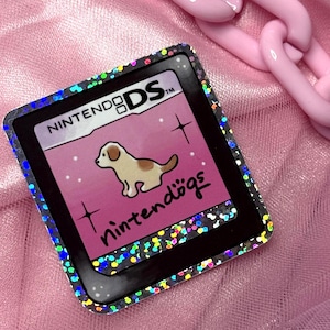 May include: A square sticker featuring the Nintendogs game logo with a light brown puppy illustration on a pink background. The sticker has a black border with a holographic, multi-colored glitter effect and is surrounded by a light pink chain.