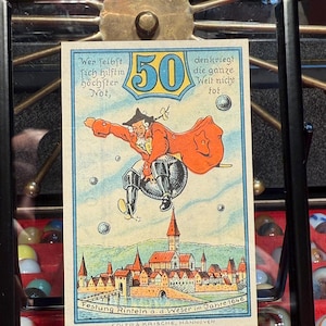 Könnte beinhalten: Gerahmter Vintage-Druck mit einer skurrilen Illustration eines Mannes in rotem Mantel und Hut, mit der Zahl 50 in einem gelb-blauen Banner. Der Druck enthält deutschen Text und eine Darstellung einer Stadt mit Fluss und Brücke. Der Text unten lautet "Festung Rinteln a. d. Weser im Jahre 1646".