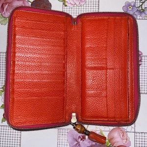Może przedstawiać: Otwarty pomarańczowy skórzany portfel z wieloma przegródkami na karty i przegródkami. Portfel ma pełne zapięcie na zamek błyskawiczny z ciemnoróżową taśmą zamka i złotym suwakiem. Wnętrze jest w żywym pomarańczowym kolorze.