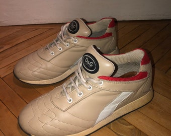 May include: A pair of beige leather trainers with red accents and a white stripe on the side. The trainers have a black logo that says "D&G" on the side.