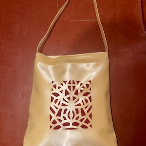 May include: A beige handbag with a single strap. The bag features a cut-out design revealing a red interior. The bag is made of a shiny material and has a rectangular shape.