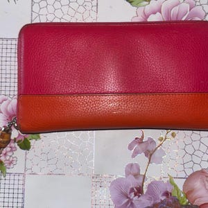 May include: A bi-color wallet in shades of red and orange. The top portion is a deep red, while the bottom is a vibrant orange. A zipper runs along three sides. A tassel with a bamboo-like accent hangs from the zipper.