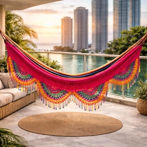 Puede incluir: Una colorida hamaca con borde de flecos, suspendida en un balcón con vistas a un paisaje urbano y agua. La hamaca presenta un centro rosa brillante con detalles y borlas de colores del arcoíris. Una alfombra redonda y tejida se encuentra debajo.