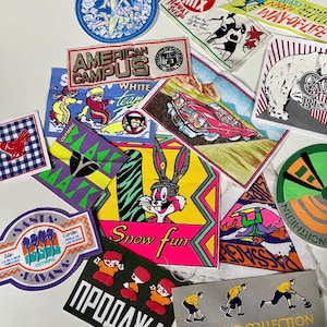 Peut inclure: Un assortiment d'écussons brodés colorés avec des motifs variés. Les écussons présentent des textes tels que "American Campus" et "Snow Fun". D'autres motifs incluent des personnages de dessins animés et une voiture vintage.