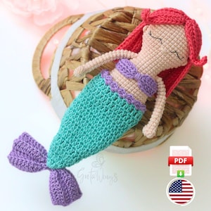 May include: A handmade crochet mermaid doll with red hair, a purple top, and a turquoise tail. The doll is lying on a woven basket. The doll is a soft toy, perfect for children.