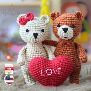 Puede incluir: Dos osos de peluche de ganchillo, uno color crema con un lazo rojo y el otro marrón, están juntos. Abrazan un corazón rojo con la palabra "LOVE" bordada. Los osos están hechos a mano y probablemente destinados a ser un regalo o un artículo decorativo.