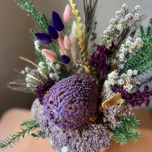 May include: A vibrant floral arrangement featuring a large, textured purple flower as the focal point. The bouquet includes accents of pink, yellow, green, and white dried flowers and foliage, creating a colorful and natural display. The arrangement is in a white vase.
