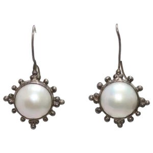 May include: A pair of silver drop earrings featuring a central round white pearl. The metal setting surrounding each pearl has a sunburst design with small silver spheres along the outer edge. Each earring hangs from a curved silver ear wire hook.