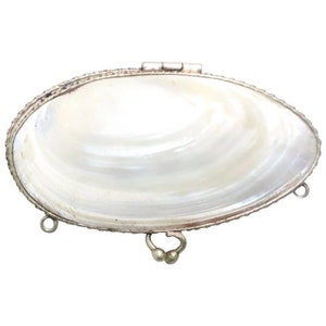 May include: A silver-toned oval-shaped trinket box with a mother-of-pearl inlay. The box has a hinged lid and three decorative loops on the bottom.