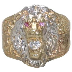 Könnte beinhalten: Ein gold- und silberfarbener Ring mit einem skulptierten Löwenkopf-Design. Der Löwe hat zwei kleine rosa Edelsteinaugen und einen klaren, rund geschliffenen Edelstein im geöffneten Maul. Die Ringschiene zeigt komplizierte, geprägte Verzierungen.