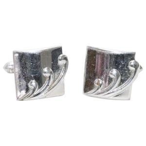 May include: A pair of square silver-toned cufflinks featuring a polished finish and a raised, curved decorative pattern on the front surface. Each cufflink has a metallic construction with a smooth, reflective face and a distinct, sweeping design.