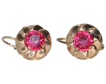 May include: A pair of gold earrings with a pink gemstone in the center of each earring. The earrings have a twisted design around the gemstone.