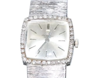 May include: A silver square-shaped wristwatch with a diamond-encrusted bezel. The watch face is white with black Roman numeral hour markers and the text "Jules Jurgensen Est. 1723" in black. The watch has a silver metal band.