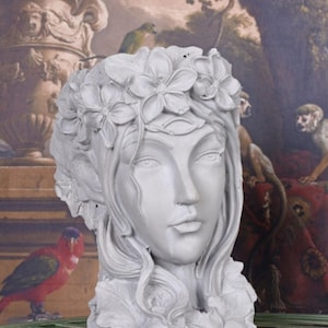 Può includere: Un vaso da fiori in cemento grigio a forma di testa di donna con una corona di fiori. Il vaso ha un viso dettagliato con gli occhi chiusi e un delicato motivo floreale intorno al collo.