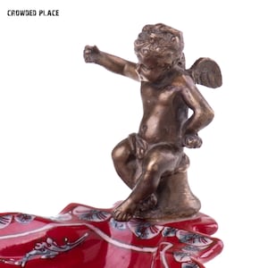 May include: A red ceramic dish with a gold cherub figurine on top. The cherub is sitting on a pedestal and has wings. The dish has a floral design.