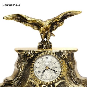 Può includere: Un orologio dorato con un'aquila appollaiata in cima. Il quadrante dell'orologio ha numeri romani e uno sfondo bianco. L'orologio è su una base decorativa con dettagli intricati.
