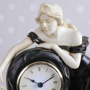 May include: A white porcelain figurine of a woman leaning on a black clock with Roman numeral markings. The clock face has gold accents and the hands are black.