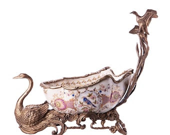 May include: A decorative bowl with a swan and bird design. The bowl is made of white porcelain with gold accents and floral patterns. The base is made of gold metal and has a swan design.